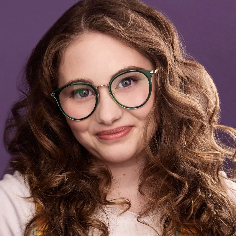 A young person with long, wavy brown hair and fair skin smiles gently. They wear round, green-rimmed glasses and a white top. The background is a solid purple color, highlighting their face and features.