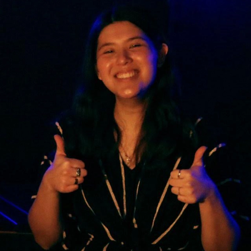 A person with long dark hair smiles broadly and gives two thumbs up. They are wearing a dark shirt with light stripes. The background is dimly lit with blue and purple lighting, creating a cheerful and positive atmosphere.