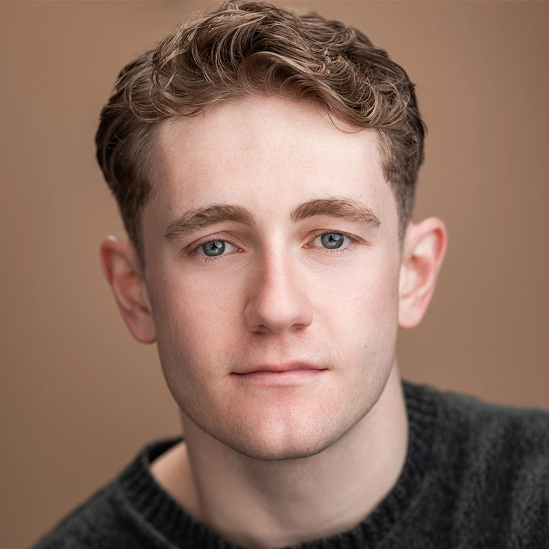 A young man with short, wavy light brown hair and blue eyes looks directly at the camera with a neutral expression. He wears a dark, textured sweater, and the background is a plain, warm brown color.