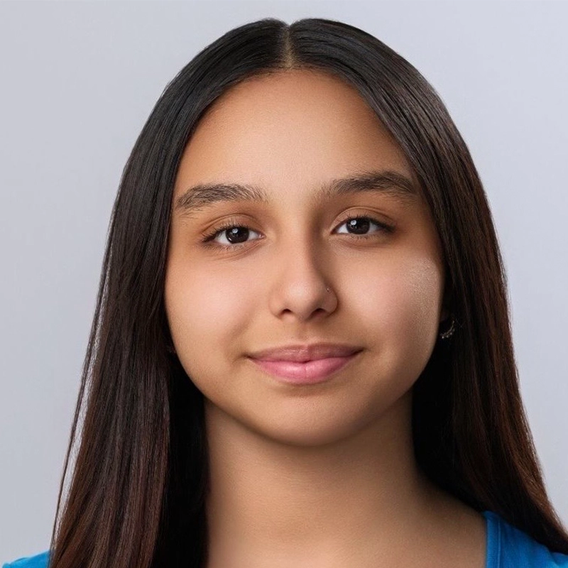 Vita Augugliaro, a young person with straight, long dark brown hair and medium skin tone, faces the camera. They have brown eyes, full eyebrows, and a gentle closed-mouth smile. They wear a blue shirt against a plain light gray background.