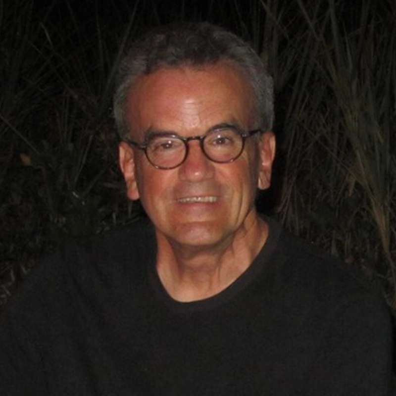 A middle-aged man with short gray hair and round glasses smiles at the camera. Peter von Mayrhauser is wearing a dark shirt and sits outdoors at night, with tall grass and plants visible in the dimly lit background.