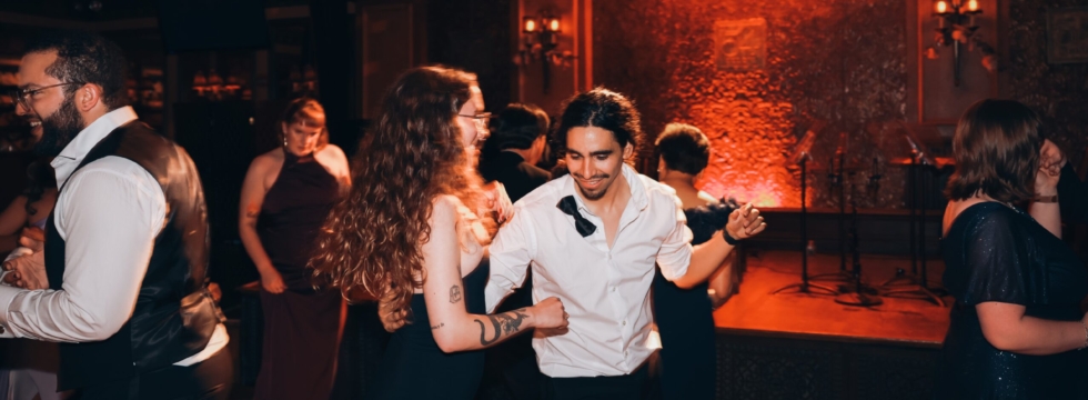 A group of people dressed formally are dancing and smiling in a warmly lit room with ornate wallpaper and chandeliers. The focus is on a man in a white shirt and a woman with long curly hair and glasses enjoying themselves.