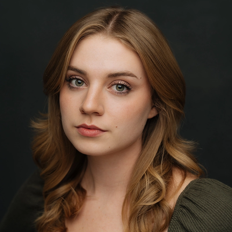 A young woman with wavy light brown hair and green eyes looks calmly at the camera. Taylor Leccese is wearing a dark green top and is posed against a dark, neutral background. Her expression is neutral and her makeup is natural.