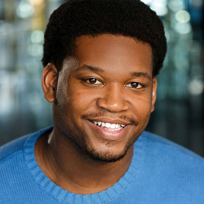 A smiling man with short curly hair and a trimmed beard wears a blue sweater. Taylor Leccese is facing the camera against a blurred indoor background with blue and gold tones, giving the image a warm and friendly feel.