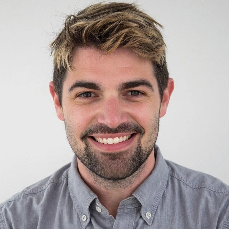 A young man with light skin, short brown hair with blonde highlights, and a trimmed beard smiles at the camera. Alex Mogil Cooke wears a gray button-up shirt and stands against a plain white background.