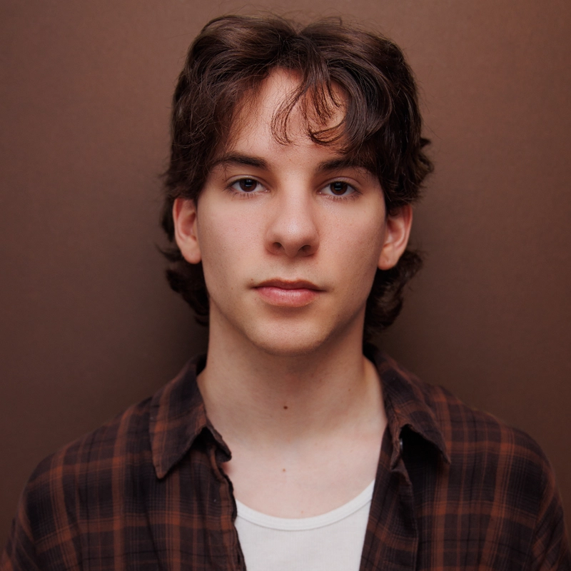 A young person with medium-length brown hair and fair skin stands in front of a plain brown background. They wear a brown plaid shirt over a white T-shirt and look directly at the camera with a neutral expression.
