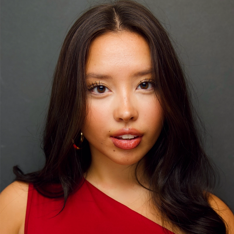 Aubrie Ofner, a young woman with long, wavy dark hair, poses against a plain dark background. She wears a red, one-shoulder top and gold hoop earrings, her neutral expression complemented by glossy lips and subtle eye makeup.