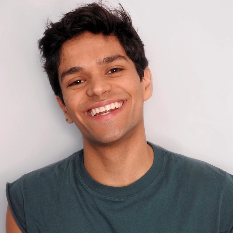 Miguel Amell, a young person with short, dark, tousled hair, smiles widely, showing teeth. They wear a sleeveless dark gray shirt against a plain light gray background. The bright lighting creates a cheerful and relaxed atmosphere.