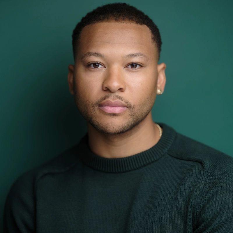 Aaron J. Anderson, a man with short, curly hair, a trimmed beard, and small gold earrings, wears a dark green crew-neck sweater as he poses calmly against a smooth, dark green background, looking directly at the camera.