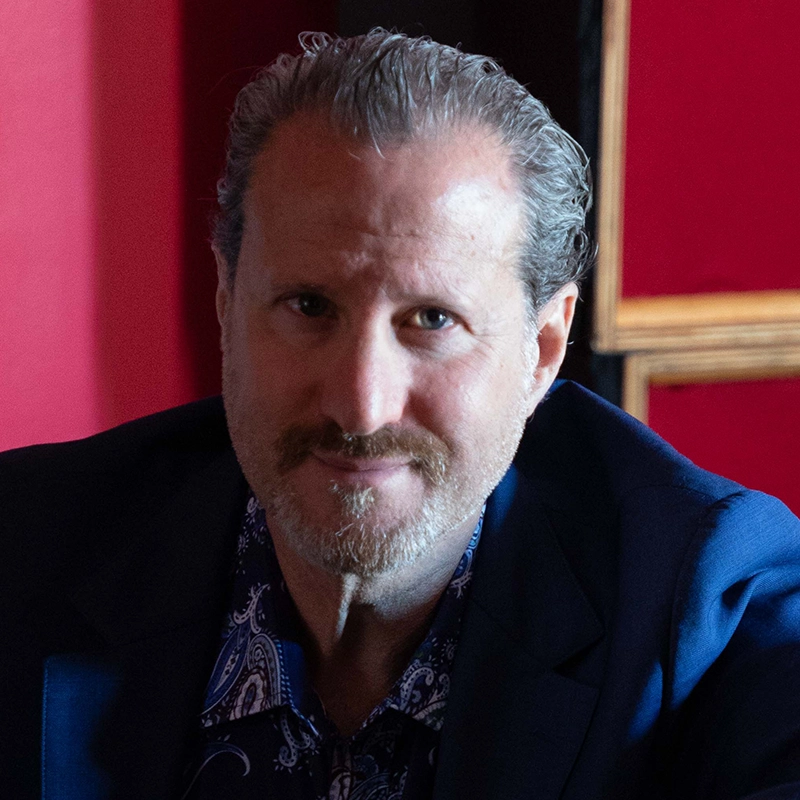 A middle-aged man with short, slicked-back gray hair and a trimmed beard sits indoors against a red wall. He wears a blue jacket over a patterned shirt and looks directly at the camera with a slight smile, illuminated by soft light from the left.