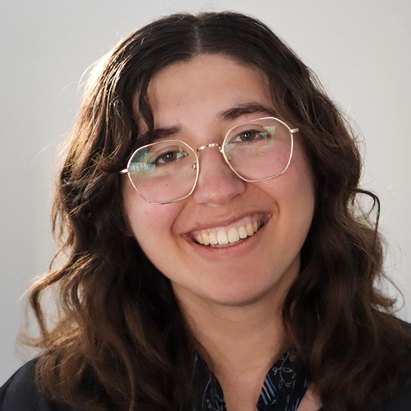 A young person with long wavy brown hair and glasses smiles warmly at the camera. They are wearing a dark jacket and a patterned scarf. The background is plain and softly lit, keeping the focus on their friendly expression.
