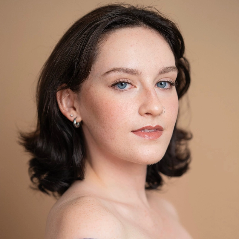 A young woman, Casey Martin Klein, with short, wavy dark brown hair poses against a beige background. She has light skin, blue eyes, and a subtle natural makeup look. She wears small hoop earrings and a faint smile, with bare shoulders visible.