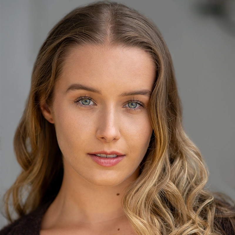 A young woman with wavy blonde hair and blue eyes looks directly at the camera. She has fair skin, subtle makeup, and a neutral expression. Captured by Michael Raul Perez, the softly blurred background draws focus to her face.