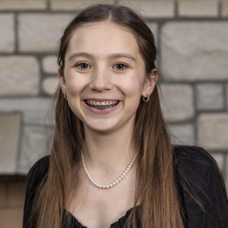 A young girl with straight, long brown hair smiles, showing braces. She wears a black top, a pearl necklace, small hoop earrings, and stands in front of a stone wall background. The lighting is warm and even.