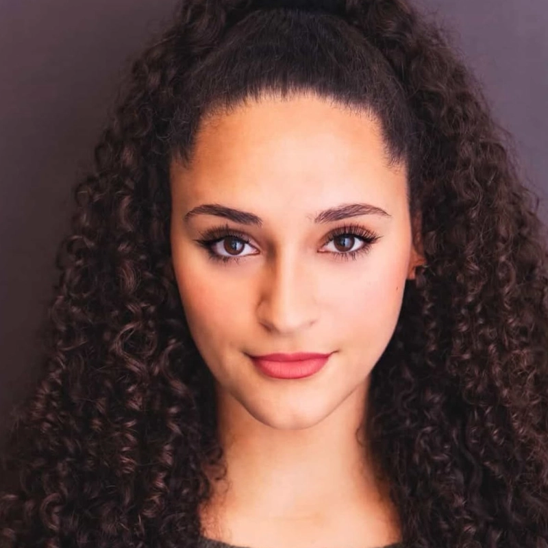 A young woman with long, dark, tightly curled hair styled in a high ponytail. She has medium skin, defined eyebrows, and brown eyes with subtle eyeliner—Hayden Barsamian faces forward against a dark background, softly smiling.