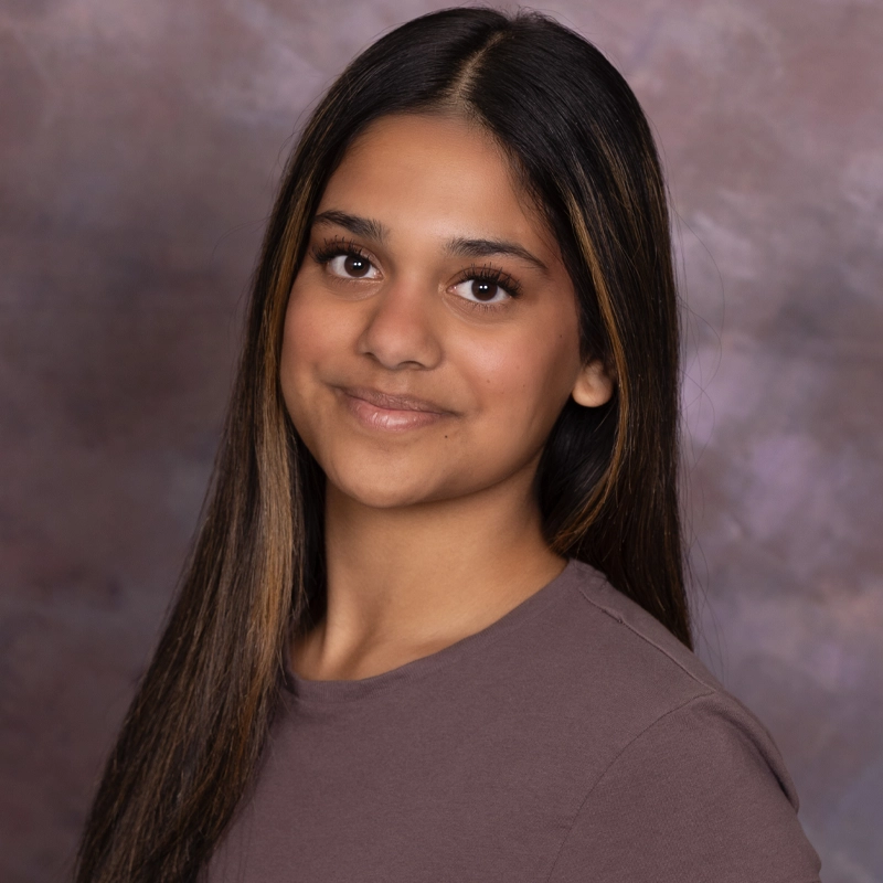 Maya Batavia, a young woman with long, straight brown hair and medium skin tone, stands in front of a soft, purple-gray mottled backdrop. She wears a mauve top and smiles gently at the camera with a relaxed, confident expression.