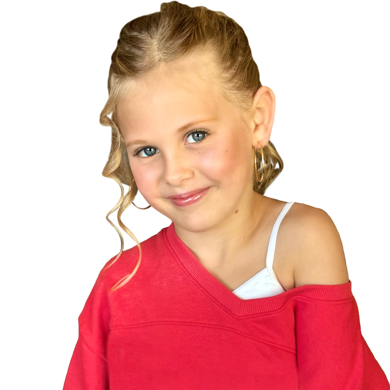 A young girl with blonde curls, resembling Alledria Bauman, smiles softly. She wears a red off-the-shoulder top with a white strap showing underneath and large hoop earrings. Blue eyes and rosy cheeks stand out against the plain white background.