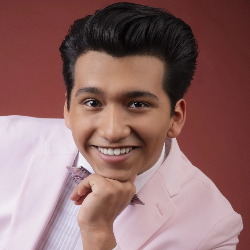 A young man with dark, wavy hair smiles warmly at the camera. Lucas Beltran wears a pale pink suit and bow tie, his chin resting on his hand. The background is a solid, muted red color. His expression is friendly and approachable.
