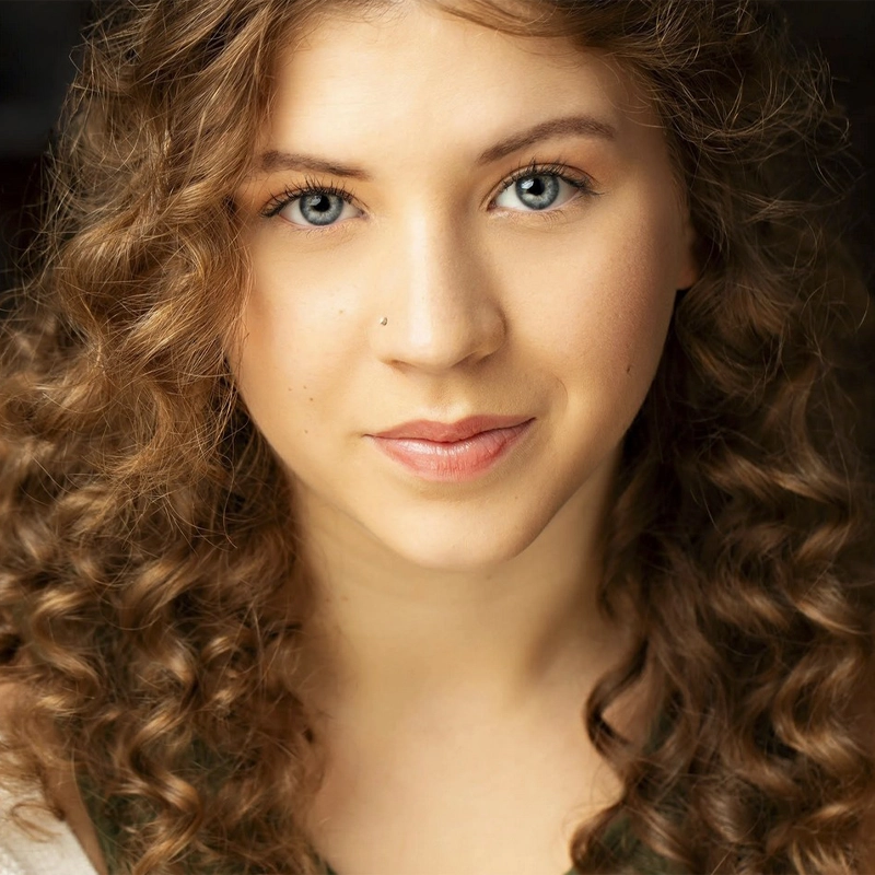 A young woman with light skin and long, curly brown hair looks directly at the camera. Brooke Benedetti has blue eyes, a small nose piercing, and a slight, closed-mouth smile. The dark background makes her brightly illuminated face stand out.