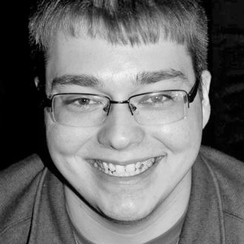 A young person smiles broadly at the camera. They have short, straight hair with bangs, wear rectangular glasses, and have visible dimples. The image is in black and white, and the background is dark, making the face the focal point.