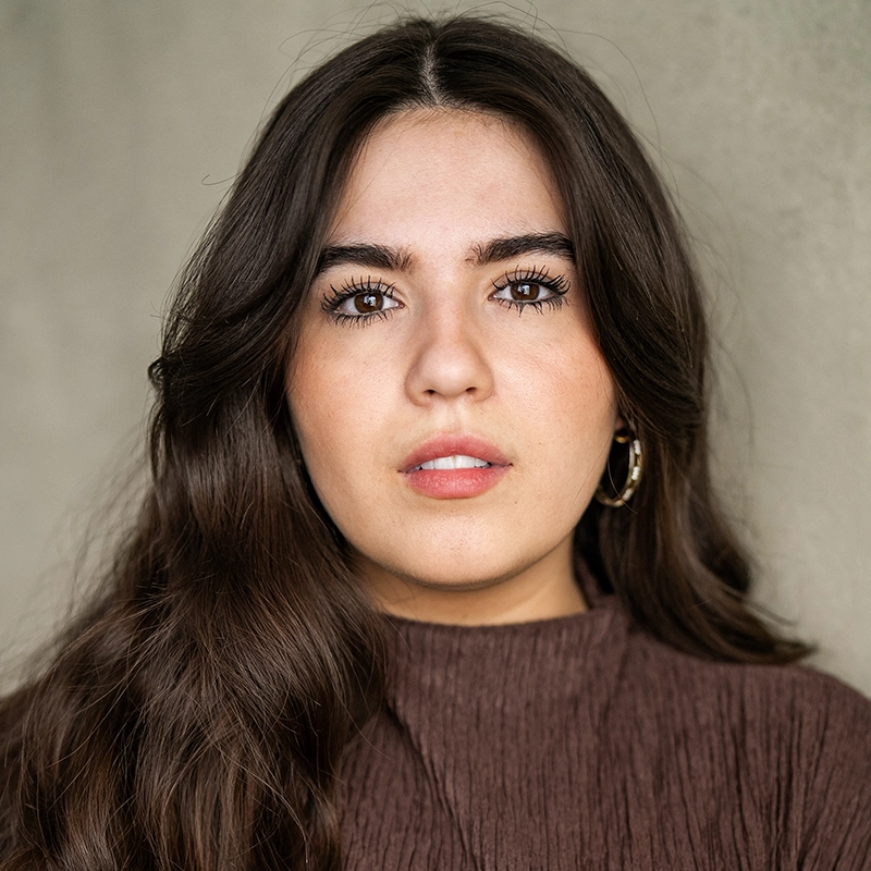 A young woman with long brown hair and brown eyes looks directly at the camera. Taylor Leccese wears gold hoop earrings and a textured brown top. The background is a plain, light-colored wall, and her expression is neutral and calm.