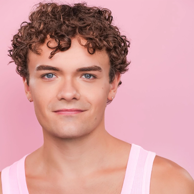A young person with short, curly brown hair and blue eyes smiles slightly. They wear a light pink tank top and a small hoop earring. The background is solid light pink, giving the image a soft, cheerful vibe.