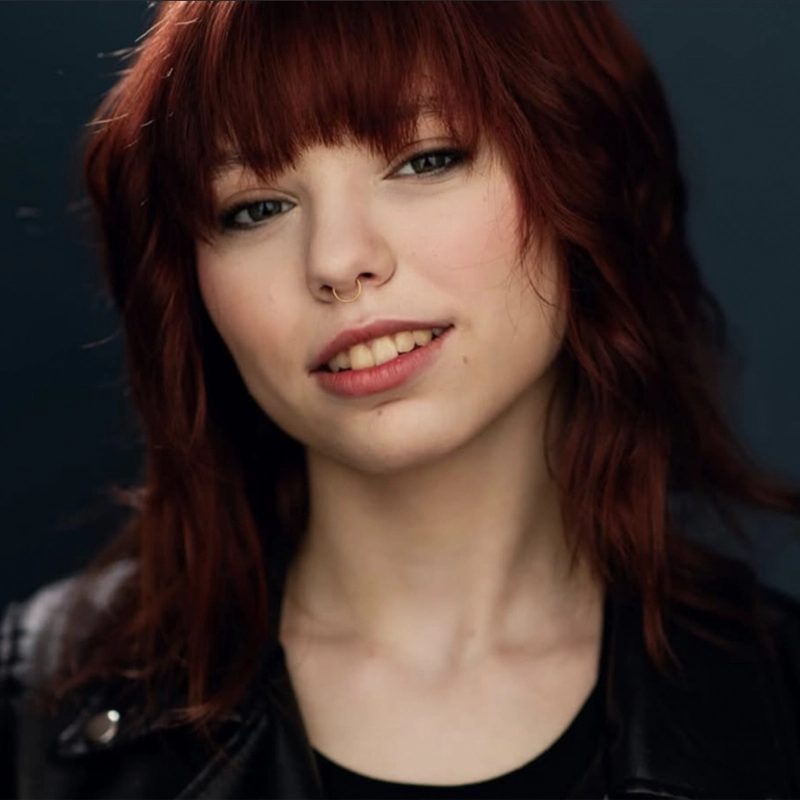 A young woman with wavy reddish-brown hair and bangs smiles softly. She has light skin, a nose ring, and wears subtle makeup. She is dressed in a black jacket and shirt, posing against a dark, blurred background.