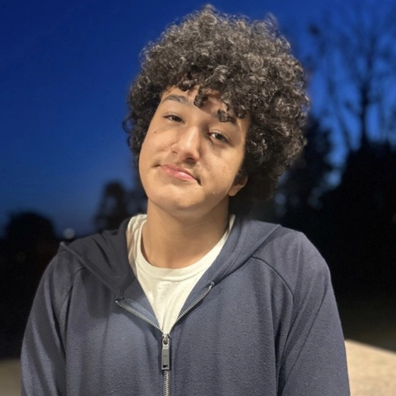 A young person with curly hair and medium skin, resembling Rayan Bouhadja, wears a dark zip-up hoodie over a white shirt, standing outdoors at dusk. Their expression is neutral, framed by a deep blue sky and silhouetted trees.