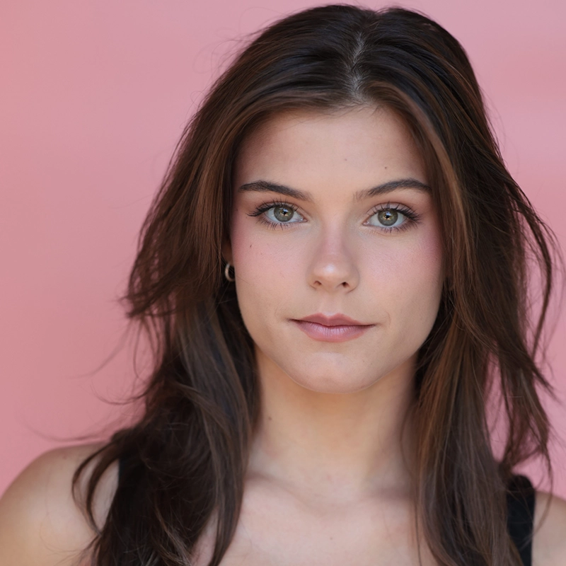 A young woman with long brown hair and green eyes, resembling Taylor Leccese, looks directly at the camera. She has natural makeup with pink blush and lipstick, and wears a black top against a solid light pink background.