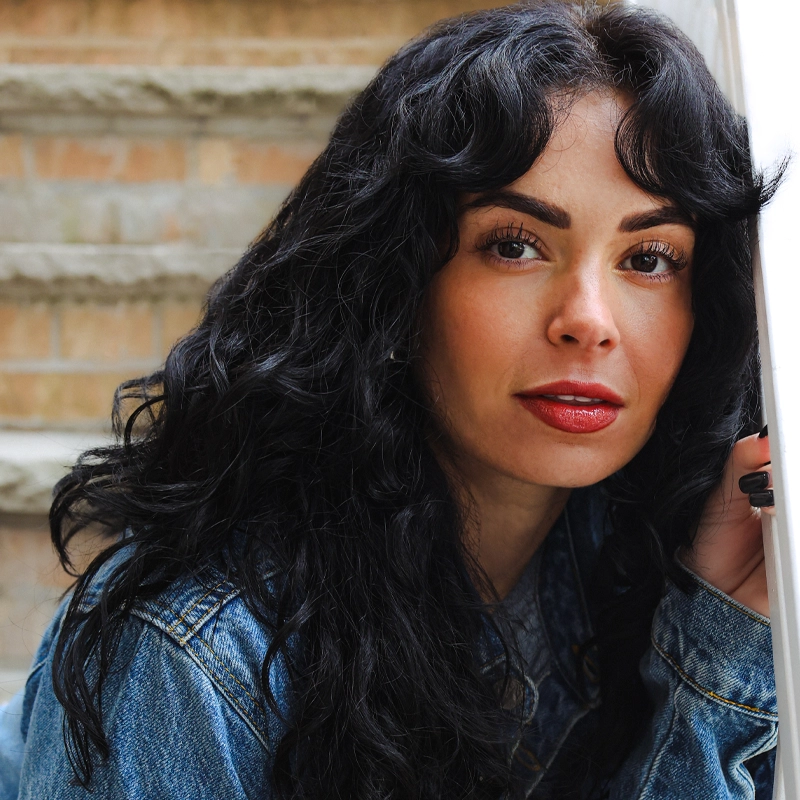 A woman with long, wavy black hair and bold red lipstick leans against a white railing. Brianna Buckner wears a blue denim jacket and looks directly at the camera with a gentle expression, framed by concrete steps and a brick wall behind her.