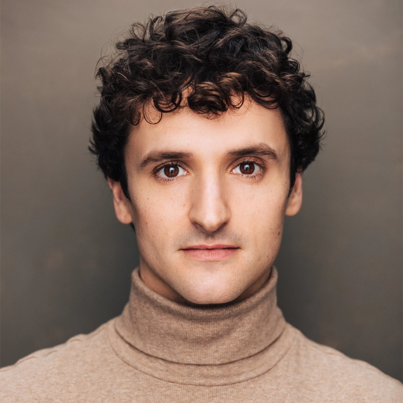 A young man with short, curly brown hair and wide brown eyes stares directly at the camera. He has fair skin and a neutral expression. Michael Bullard is wearing a light brown turtleneck sweater against a plain, muted brown background.