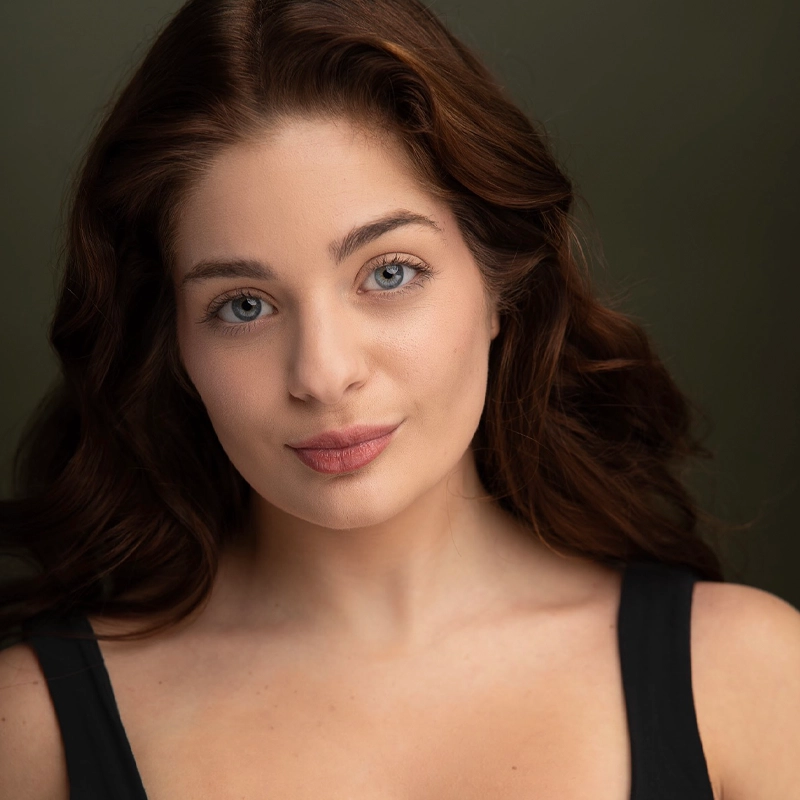 Kinsey Gray Calderone, a young woman with long, wavy brown hair and blue eyes, looks at the camera with a gentle smile. She wears a sleeveless black top against a muted, dark olive green background, softly lit to highlight her face.