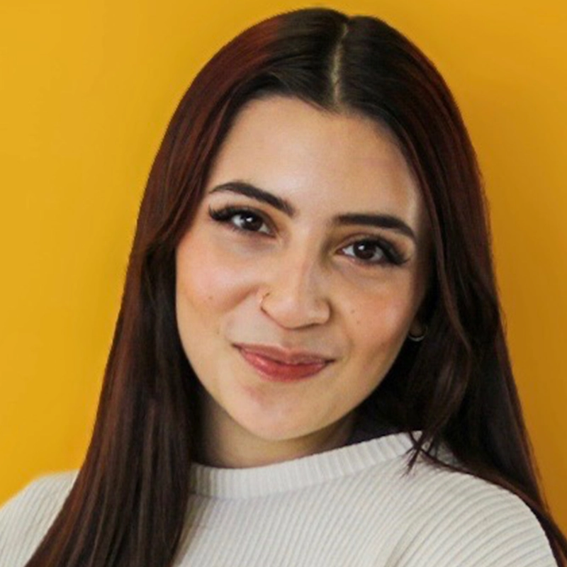 Carmelina Glynn, a young woman with long, straight brown hair and brown eyes, smiles gently. She wears a white ribbed top and subtle makeup. The vibrant yellow background creates a warm and cheerful atmosphere.