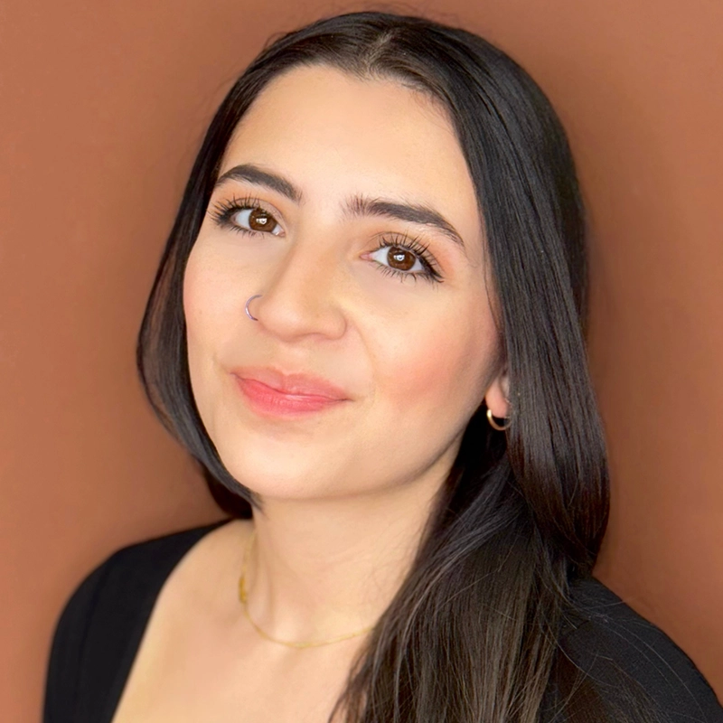 A woman with long, straight dark hair and light skin smiles softly at the camera. She has brown eyes, natural makeup, and wears a black top, a small nose ring, hoop earrings, and a delicate gold necklace. The background is solid brown.