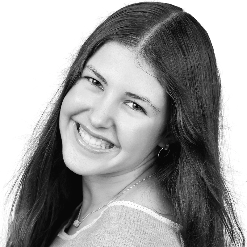 A young woman with long, dark hair smiles warmly at the camera. Grace Carnicelli has light skin and wears a necklace, hoop earrings, and a light-colored top. The black-and-white portrait features a plain white background.