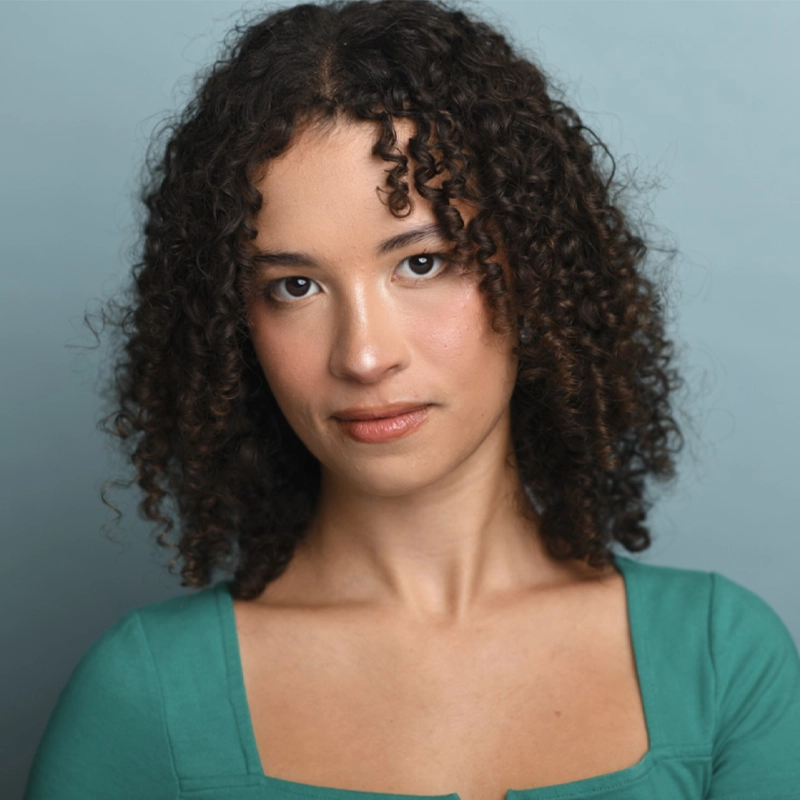 Holly Carter Berman, with medium brown curly hair and light skin, gazes confidently at the camera. She wears a teal square-neck top and natural makeup against a soft, solid light blue background for a calm, professional portrait setting.