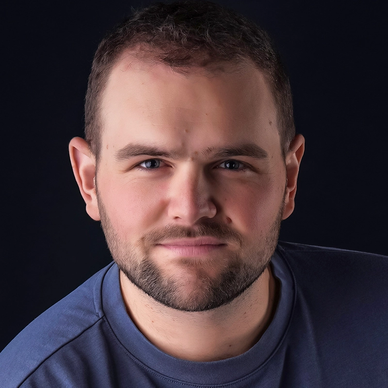 Casey Martin Klein, a man with short brown hair and a beard, smiles softly at the camera. He has light skin, blue eyes, and wears a blue crewneck shirt. The solid black background gives the portrait a professional and focused appearance.