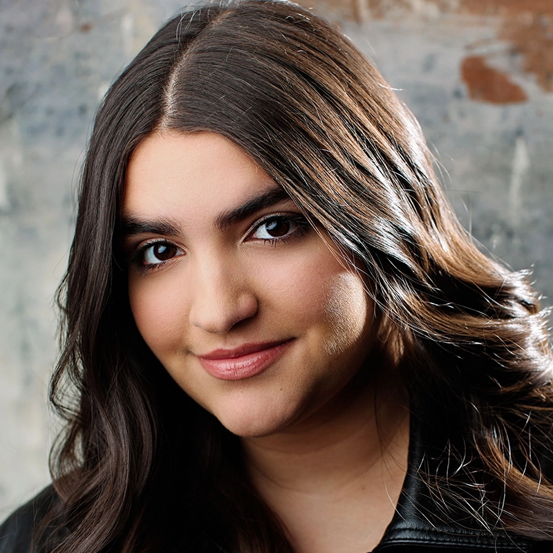 Aubrie Ofner, a young woman with long, wavy dark hair, smooth skin, and brown eyes, smiles gently. She wears a black jacket and stands before a textured grayish background accented with brown and white. The lighting highlights her face and hair.