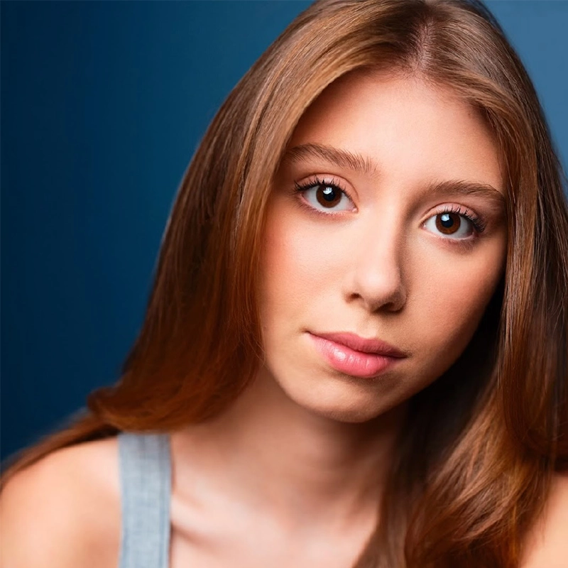 A young woman with long straight auburn hair and brown eyes looks directly at the camera. She has fair skin, natural makeup, and wears a gray tank top. The background is a gradient of dark to light blue.