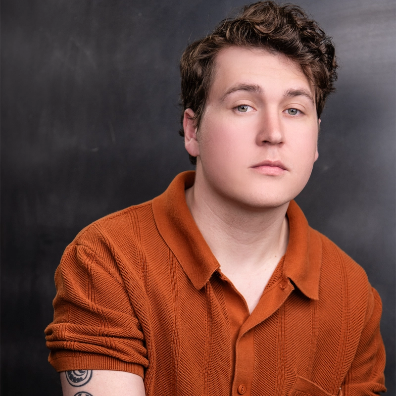 A young person with short, wavy brown hair wears an orange textured polo shirt. They have light skin, subtle makeup, and a serious expression. A tattoo is visible on their upper left arm. The dark, blurred backdrop highlights Ryan J. Charest's striking presence.