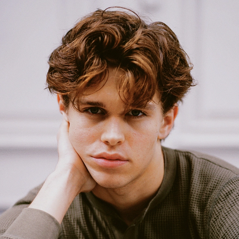 Aubrie Ofner, a young person with wavy brown hair and fair skin, rests their face on their hand, looking directly at the camera with a neutral expression. They wear a textured olive-green top against a softly blurred light gray background.