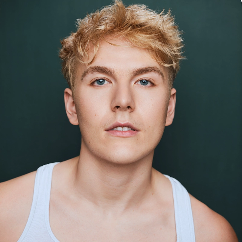 Casey Martin Klein, a young person with short, tousled blond hair and blue eyes, looks directly at the camera. They wear a white tank top and have a neutral expression. The background is solid dark green.