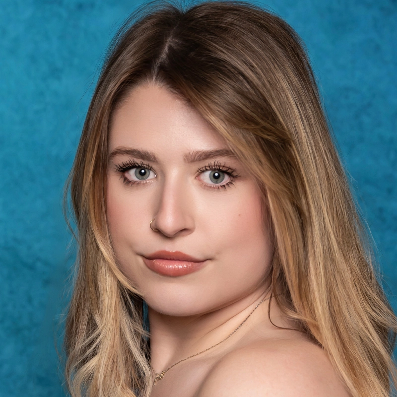 A young woman with long, straight blonde hair poses in front of a bright blue textured background. Megan Culpepper, with light skin and blue eyes, wears natural makeup and nude lipstick, gazing directly at the camera with a composed expression.