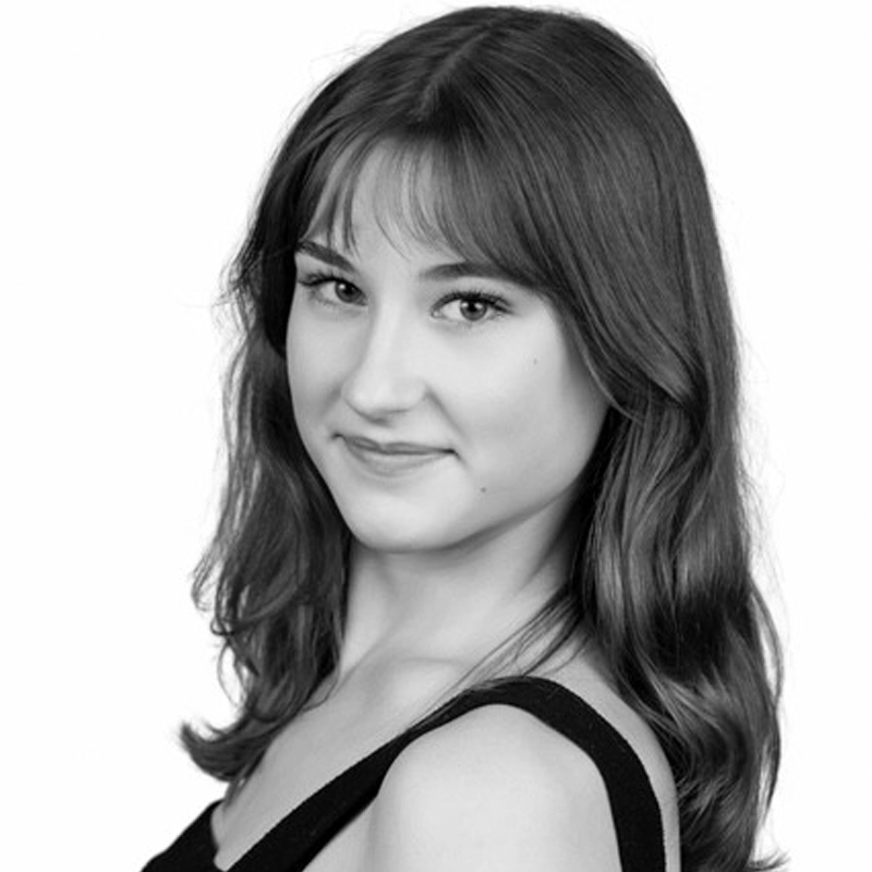 Black and white portrait of Maggie Cummings, a young woman with wavy, shoulder-length hair and bangs. She smiles gently over her shoulder at the camera, wearing a sleeveless top against a plain white background.