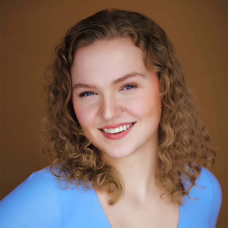 A young woman with curly, light brown hair smiles warmly at the camera. She wears a light blue top and has a small nose piercing. The background is a soft, warm brown, and the lighting highlights her clear skin and blue eyes.