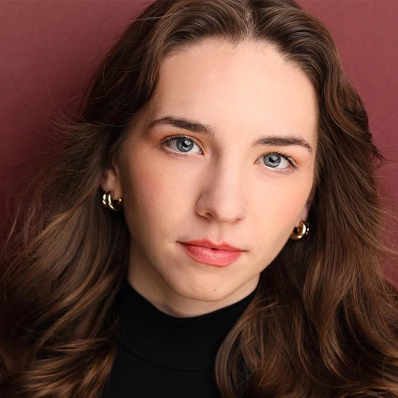 A young woman with wavy brown hair and blue eyes looks directly at the camera. She wears gold hoop earrings, a black top, and natural makeup. The background is a solid, muted maroon color. Her expression is calm and neutral.