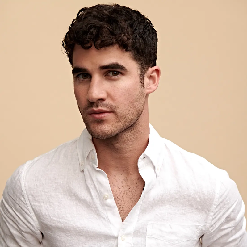 Darren Criss, wears a white linen button-up shirt. He looks directly at the camera with a neutral expression. The background is plain beige with soft, even lighting.
