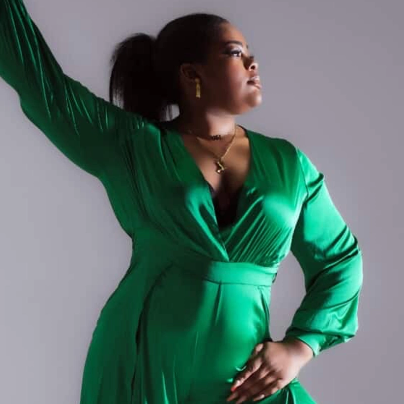 A woman wearing a vibrant green, long-sleeve, wrap dress poses confidently against a plain background. She has her hair in a ponytail, wears gold jewelry, and looks off to the side with one arm raised, exuding strength and elegance.