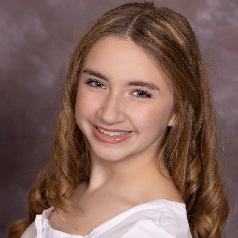 A smiling young woman with long, wavy light brown hair poses in front of a soft, mottled brown and gray background. She wears a white top and has natural makeup, with visible eyelashes and glossed lips. She is facing slightly to the left, looking at the camera.