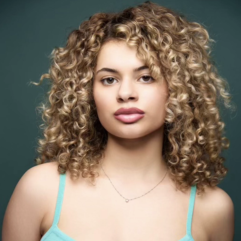 A young woman, Grace Del Corral, with curly, light brown hair and highlighted blonde strands poses against a dark green background. She wears a turquoise spaghetti strap top and a delicate necklace, looking directly at the camera with a neutral expression.